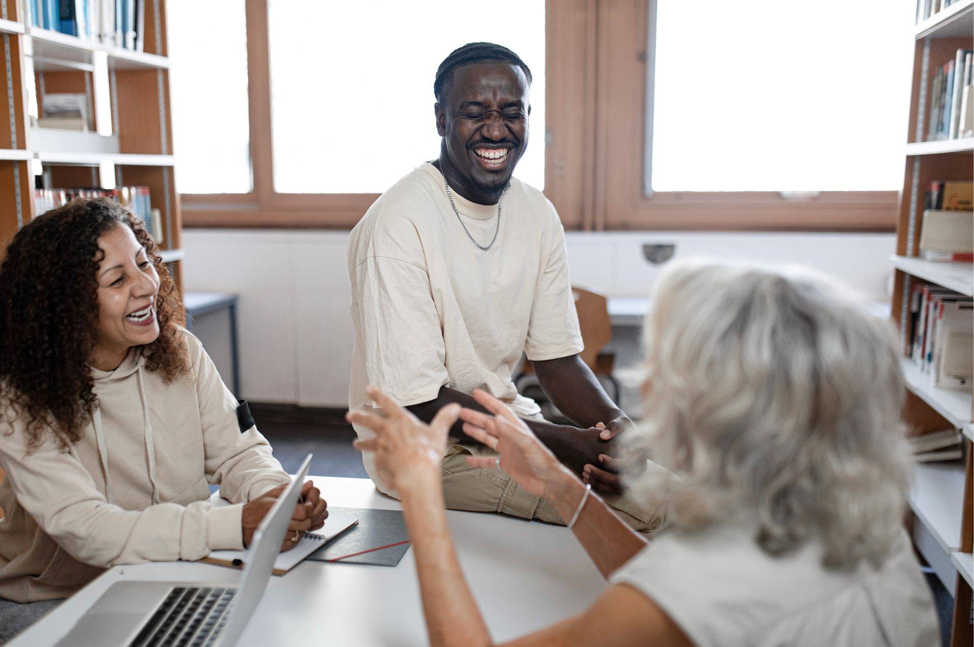 A man is sat on a desk talking to two women. They're laughing and have laptops on the desk.